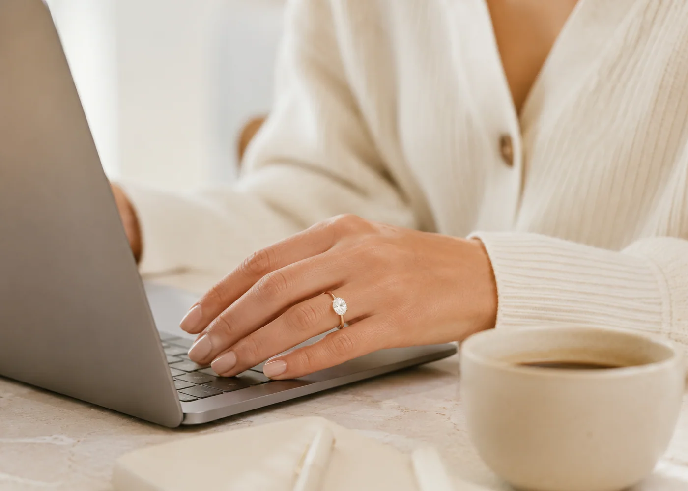 Woman wearing an engagement ring while working on a laptop beside a coffee cup.